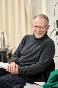 Photo from the rehearsal room. Danny Webb (grey hair brushed back, white bear, clear glasses and a roll neck) is sat on a chair hands in his lap and holding a script. He's looking off and smiling gently.