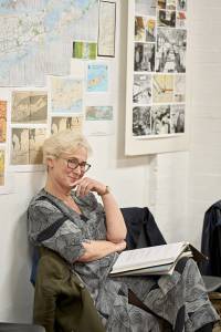 Photo from the rehearsal room. Helen Schlesinger (white hair which is short and wavy, and tortoishell glasses) is sat on a chair and leaning back against a white brick wall. She has an open script on her lap and has one hand to her chin as she smiles. On the wall behind her are maps of New York and old black and white photographs.