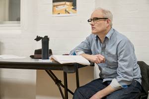 Photo from the rehearsal room. Ian Kirkby is sat side on to a long table. He has dark framed glasses and a blue collared shirt and he's looking off as if listening intently. He has one arm leaning on the table and one in his lap. On the table is an open script, a metal water bottle and a trilby hat.