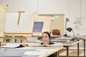 Photo from the rehearsal room. The room is full of desks and display boards covered in blue prints and buts of paper. Most of it is out of focus apart from a woman (Siobhán Cullen, brown hair pull back in a lose ponytail) who is kneeled behind one of the tables, her arms resting on the top of it and her chin resting on her knuckles. She has an open mouth smile and is looking off into the distance.