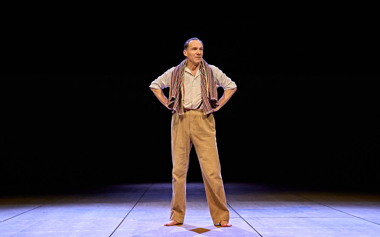 Ralph Fiennes stands centre stage, with darkness behind him. His hands are on his hips and he looks off to the side. He's wearing a white striped shirt with braces connected to the top of his trousers. There's a red and white striped towel hung around his neck.