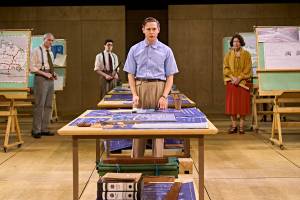 Samuel Barnett stands as the focus of the image in a blue shirt and brown smart trousers. He stands behind a big table with deign plans atop them. In the background are presentation boards with other architectural plans on.