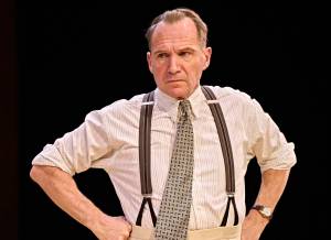 Ralph Fiennes stands with hands on his hips with his brows furrowed. He's wearing a white striped shirt, tie and braces.