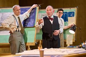 Ian Kirby stands leaning on a wooden board that shows blueprints and maps. He is wearing a white striped shirt, braces and a tie. Danny Webb stands with a cigar between his fingers talking. He is wearing a pin striped waistcoat and red silk bow tie. In the background Al Coppola is watching Danny Webb. He wears a white shirt and braces.