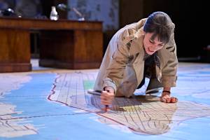 Siobhán Cullen kneels on the floor, she is drawing in red pen on a huge map of New York which covers the floor. She's wearing a trench coat and is clenching her teeth.
