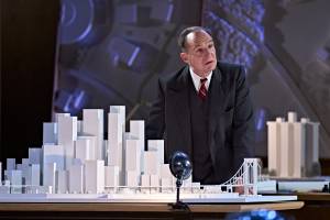 Ralph Fiennes looks pensive stood behind a 3D paper model of New York City, with sky scrappers and large buildings. He's wearing a dark suit and waistcoat and a red tie.