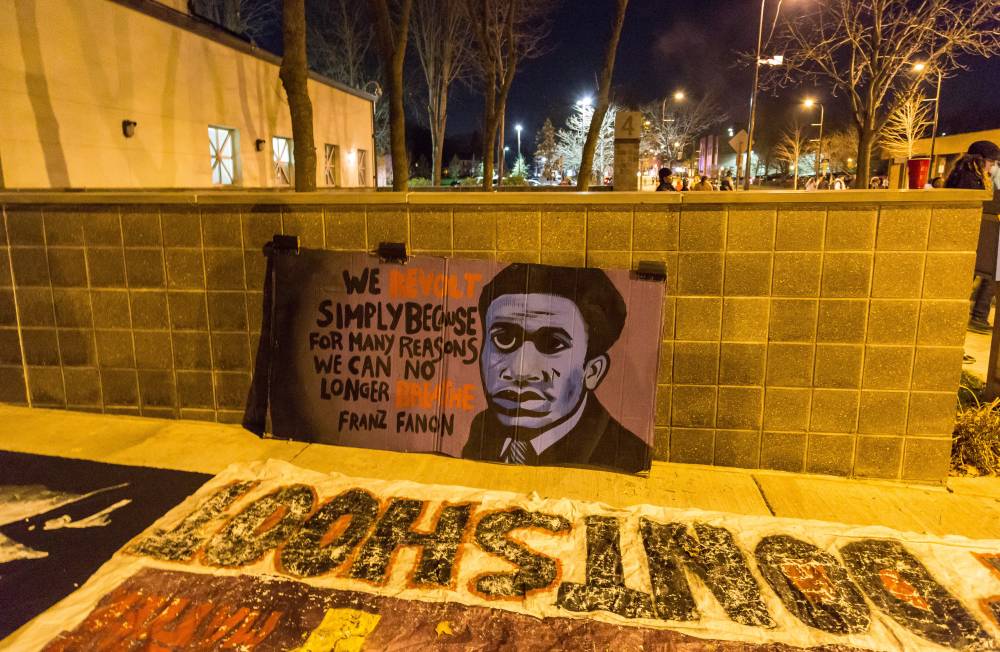 At night time, a hand-painted banner reads 'We revolt simply because for many reasons we can no longer breathe' – Franz Fanon