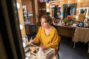 Mary Stillwaggon Stewart sits in costume at a mirror in her dressing room. She has a short brown wig and a yellow shirt.