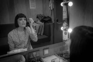 Siobhán Cullen sits at a dressing room table putting mascara on, she wears a short dark brown wig styled in a bob.