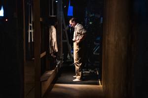 Ralph Fiennes stands backstage, with darkness around him. His hands are on his hips and he looks at the floor. He's wearing a white striped shirt and beige trousers.