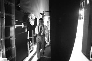 Al Coppola walks backstage smiling at the camera, he wears a shirt, trousers and braces.