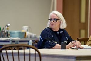 Holly Atkins sits at a wooden table leaning on a script with a pen in hand. She has white blonde hair and wears a navy top with thick black glasses.