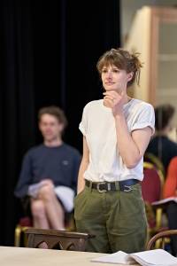 Jo Herbert stands with one hand under her chin and another in her pocket. She has brown hair tied back in a bun and wears a white t-shirt with khaki green trousers.
