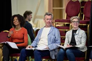 Left to right, Racheal Ofori, Alex Jennings and Hermione Gulliford sit on chairs ina row against a wall of mirrors. There attention is focused of camera, as they smile. Racheal wears a red longs sleeved top and gold jewellery, Alex has blue overshirt ontop of a white top, his glasses sit on his head. Hermione wears a black t-shirt and white denim jacket with glasses. They each hold scripts.
