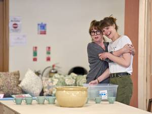 Phoebe Nichols and Jo Herbert stand behind a table with a large bowl and series of mugs atop it. Phoebe embraces Jo with a shocked expression whilst Jo laughs.