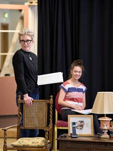 Hermione Gulliford stands with a script in one hand and the other resting on a rattan wooden chair. She's got dark framed glasses and short hair style in a quiff.