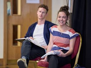 Sarah Twomey (brown long hair in a messy top knot) sits smiling. She's wearing a blue red and white striped top and has a script open resting on her knees. In the background slightly blurred is Jack Greenlees (short curls) sat in the same way.