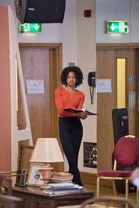 In amongst a busy room filled with household props, Racheal Ofori stands holding a script over in her arms. Her legs are slightly crossed and she looks off into the distance. She has dark hair in tight curls, and is wearing an orange long sleeved top and black leggings.