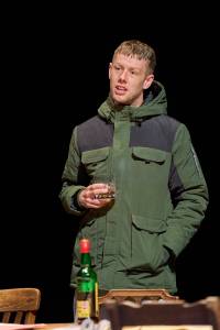 Josh Finan stands behind wooden table, he wears a green coat and holds a glass in one hand and the other in his pocket. His mouth is slightly open and looks affronted.