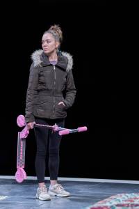 Sarah Twomey stands holding a pink children's scooter, she has a blank expression on her face and wears a puffer jacket.