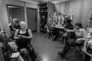 Black and white photo of a large dressing room. There are dressing tables around the edge of the room that have mirrors with lights around them, along with celebration greeting cards. 5 women from the cast sit at their tables and are all mid laughter
