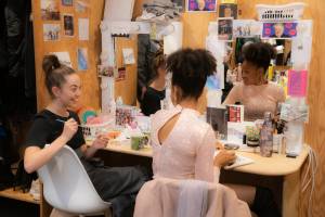 Sarah Twomey (brown long straight hair in a messy top knot) and Racheal Ofori (brown afro hair in a high pony tail) sit at their dressing room mirrors, both are busy crafting with their hands but they look at each other smiling