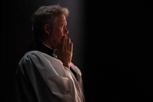 Alex Jennings (grey curly hair) is wearing a white cassock and dog collar, his hands are to his mouth