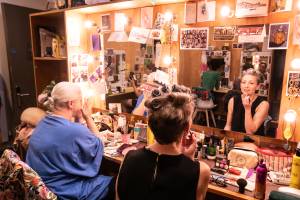 Holly Atkins (bleach blonde hair tied back in a low bun) and Hemione Gulliford (cropped blonde hair in rollers) sit in front of dressing room mirrors doing their make up. There are lights around the mirror and greetings cards all over the wall