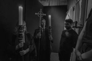 Backstage, waiting to go on are three people holding ornately decorated candlesticks and a cross.