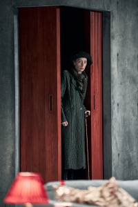 Lia Williams stands in a red doorway with a cane, she wears a long coat and a hat, her face looks wary.