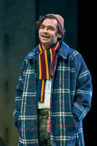 Sebastian De Souza stands with his hands in his coat pockets smiling widely. He wears a patterened winter coat, a beanie hat and a stripey scarf.