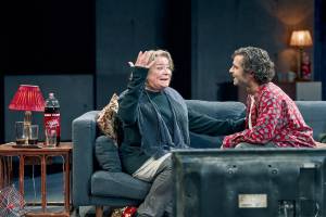 Clare Higgins and Sebastian De Souza laugh together on a sofa, Clare looks bemused as Sebastian watches her fondly.
