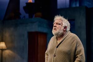 Simon Russell Beale stands looking upwards deep in though, his hair is messy and he wears an old beige cardigan.
