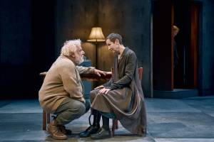 Simon Russell Beale and Lia Williams, Lia sits on a wooden chair and Simon crouches in front of her, they look at each other intently.