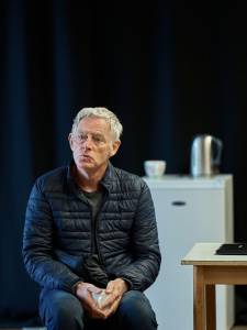 Michael Simkins perches on a stool, wearing a puffer jacket and clasping a mug in his hands. His lips are pursed together and his head cocked slightly.