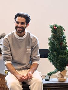 Eben Figueiredo sits on a wooden chest next to a small Christmas tree with his hands in his lap. He's smiling widely and wears a grey jumper with greige trousers.
