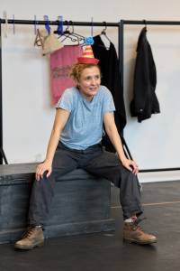 Lyndsey Marshal sits on top of a wooden trunk. She wears a striped party hat, t shirt and trousers. As she sits she rests her hands on her knees.