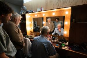 Sebastian De Souza, Simon Russell Beale and Michael Simkins getting ready backstage. They sit at dressing tables and stand in front of lit mirror.