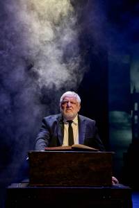 Simon Russell Beale sits behind a wooden chest wearing a suit and tie with a book in front of him and fog billowing behind.