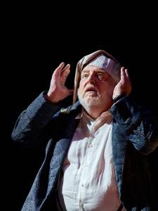 Simon Russell Beale looks upwards with his hands on either side of his face, he wars a nightcap and gown.