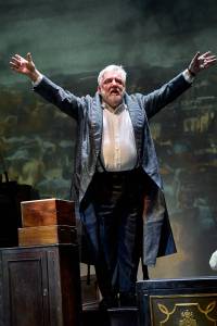 Simon Russell Beale stands with his arms outstretched wearing a long coat and white shirt.