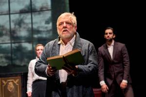 Simon Russell Beale stands with a book open in his hands looking upwards slightly.