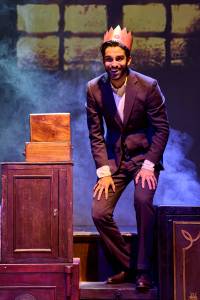 Eben Figueiredo (tall, dark hair and short beard, 20s) stands upon a stack of wooden chests. He has a paper Christmas hat on and is grinning with his hands on his knees