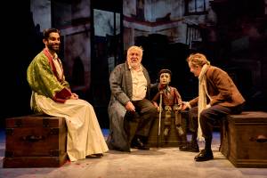 On three wooden chests sit Eben Figueiredo (in a long white apron), Simon Russell Beale in an old fashioned dressing gown and sat with a small wooden puppet of a little boy, and Lyndsey Marshal who holds the puppets other hand