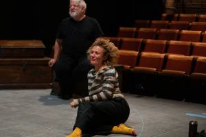 Sat on the stage of theatre sits a woman (Lyndsey Marshal), who is smiling, and a man (Simon Russell Beale) sits behind her on a wooden chest