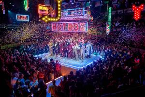 Huge crowds surround a raised platform where the cast of 28 take their bows. Confetti falls on them and neon signs hang above them