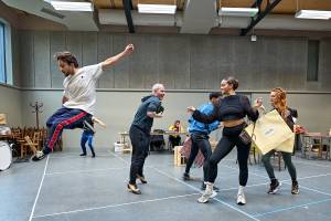Cast member dance in rehearsals, one jumps high whilst others move playfully across the room.