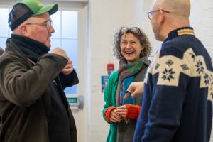 Three adults engage in a cheerful conversation in a rehearsal room with white walls.