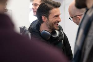 Jonathan Bailey smiles wearing a black jacket and headphones, engaged in a conversation.