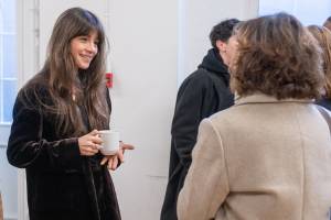 Olivia Popica holds a white mug and smiles across the room at someone facing away from the camera.